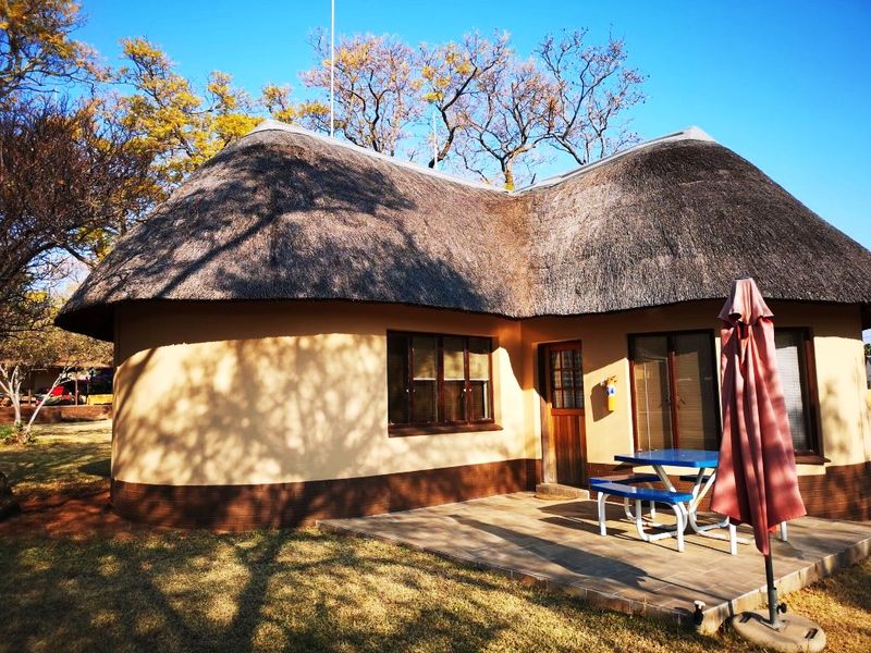 A charming lodge unit featuring a traditional thatched roof, exterior plaster finish, and a private tiled patio area set within a landscaped garden.