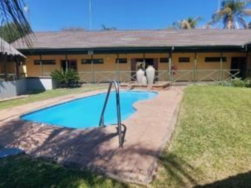 Outdoor pool area featuring a swimming pool with a ladder, paved surround, and a view of the lodge building with a thatched roof.