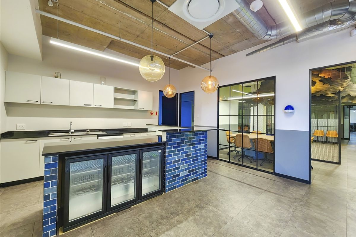 Modern office kitchenette featuring an exposed concrete ceiling, sleek cabinetry, a tiled island with integrated refrigeration, and a glass-partitioned meeting room.