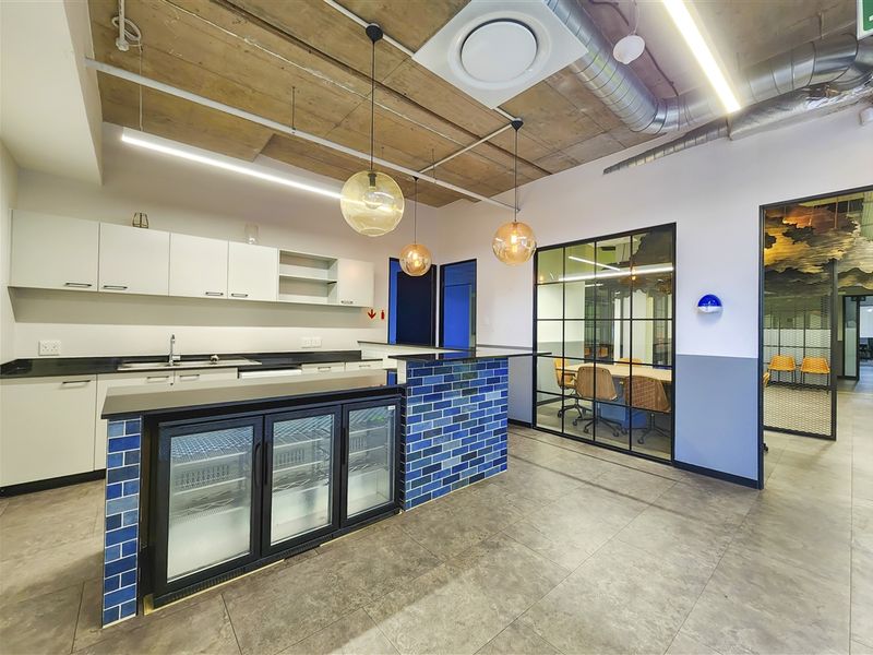 Modern office kitchenette featuring an exposed concrete ceiling, sleek cabinetry, a tiled island with integrated refrigeration, and a glass-partitioned meeting room.