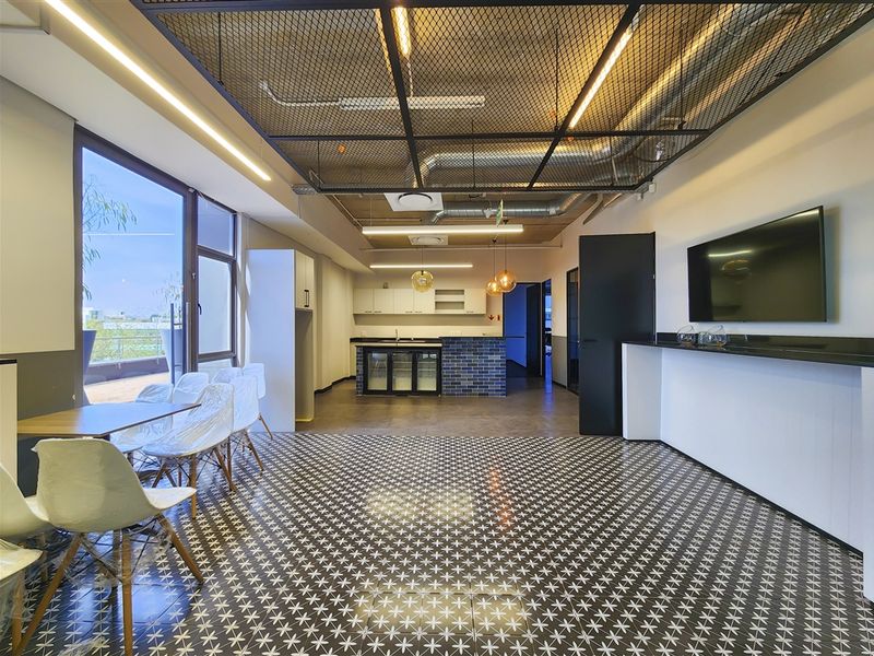 Modern office space featuring an exposed industrial ceiling, kitchenette, patterned tiled flooring, and large glass doors leading to an outdoor area.