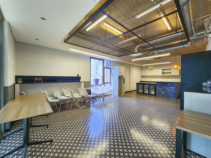 Modern open-plan office space featuring industrial-style exposed ductwork, a suspended mesh ceiling, patterned floor tiles, and a functional kitchenette area.