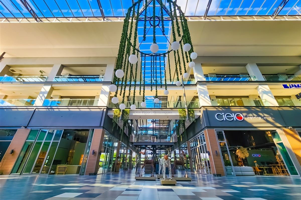 Modern commercial atrium featuring a glass roof, multi-level mezzanine with glass balustrades, and high-end storefronts with tiled flooring.