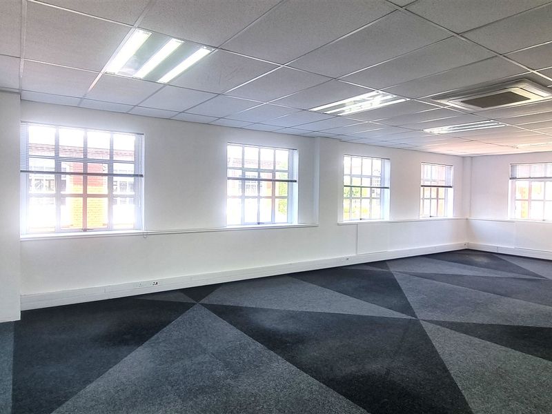 A spacious, open-plan office area featuring a suspended ceiling, recessed lighting, air conditioning, and patterned carpet tiles with ample natural light.