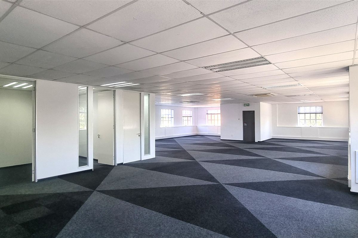 A spacious, open-plan office area featuring a suspended ceiling, recessed lighting, and patterned carpet tiles, with several partitioned internal office rooms.