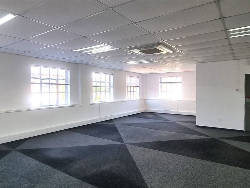 A spacious, open-plan office area featuring carpet tiles, suspended ceilings with recessed lighting, and multiple windows providing ample natural light.