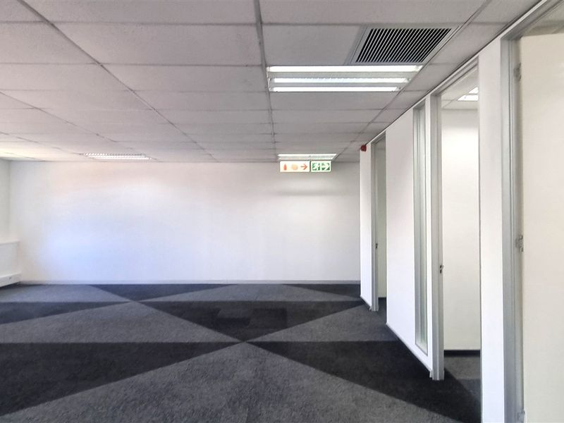 A spacious, vacant open-plan office area featuring a suspended ceiling, recessed lighting, patterned carpet tiles, and glass partition walls.
