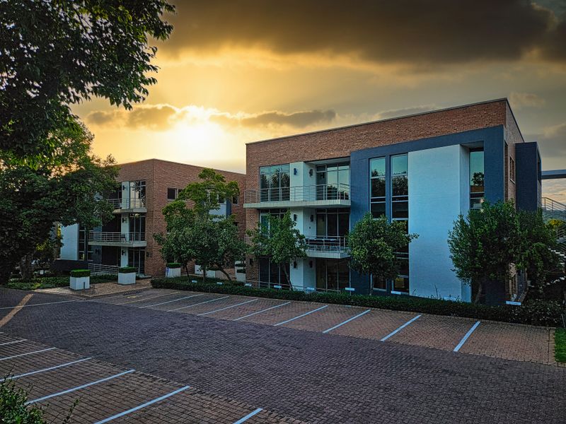Modern multi-story office building featuring brick and rendered facade, private balconies, and an extensive paved parking area at sunset.