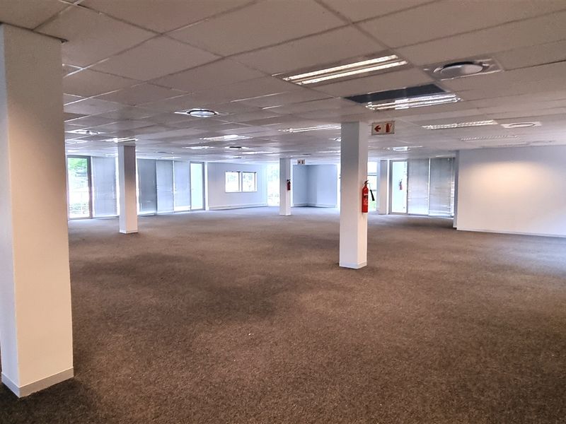 A spacious, open-plan office area featuring a suspended ceiling, recessed lighting, structural columns, and large windows with blinds, currently in a vacant state.