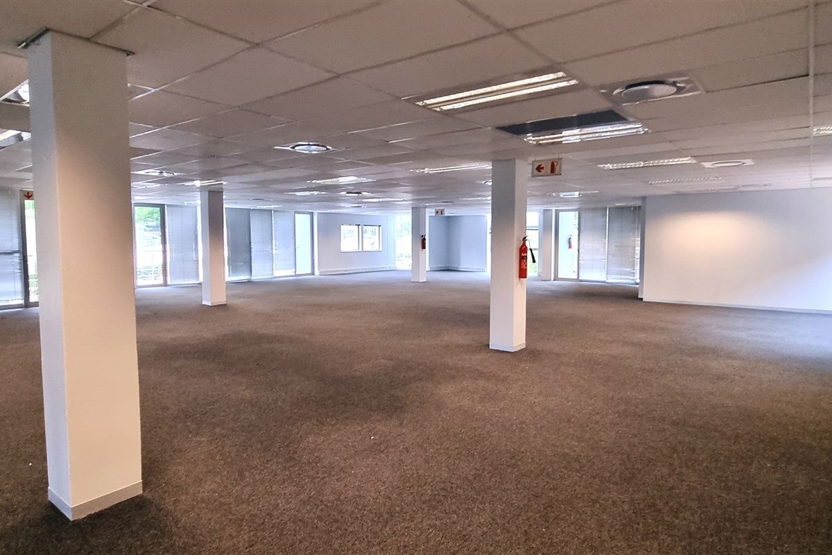 A spacious, open-plan office area featuring a suspended ceiling, recessed lighting, structural columns, and large windows with blinds, currently in a vacant state.