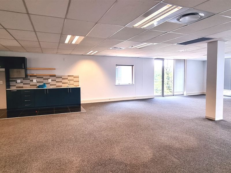 Spacious open-plan office area featuring a kitchenette, carpeted flooring, suspended ceilings with recessed lighting, and large windows providing ample natural light.