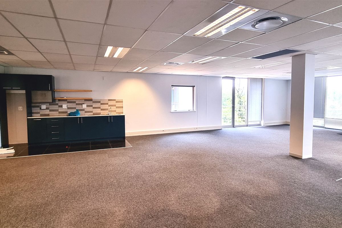 Spacious open-plan office area featuring a kitchenette, carpeted flooring, suspended ceilings with recessed lighting, and large windows providing ample natural light.