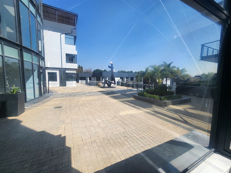 A spacious paved outdoor courtyard area featuring modern glass office architecture, raised garden beds, and metal safety railings.