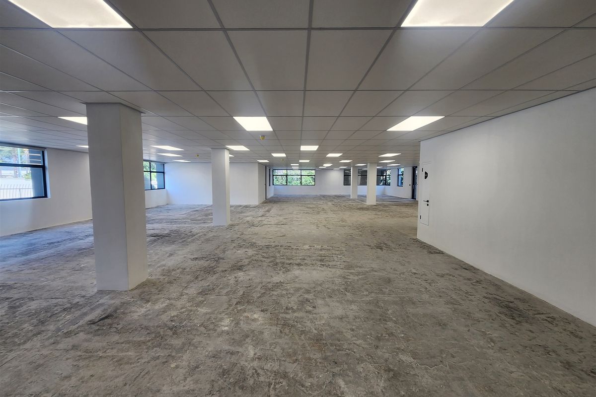 A large, open-plan office space featuring a suspended ceiling with recessed lighting, structural columns, and large windows, currently awaiting floor finishing.