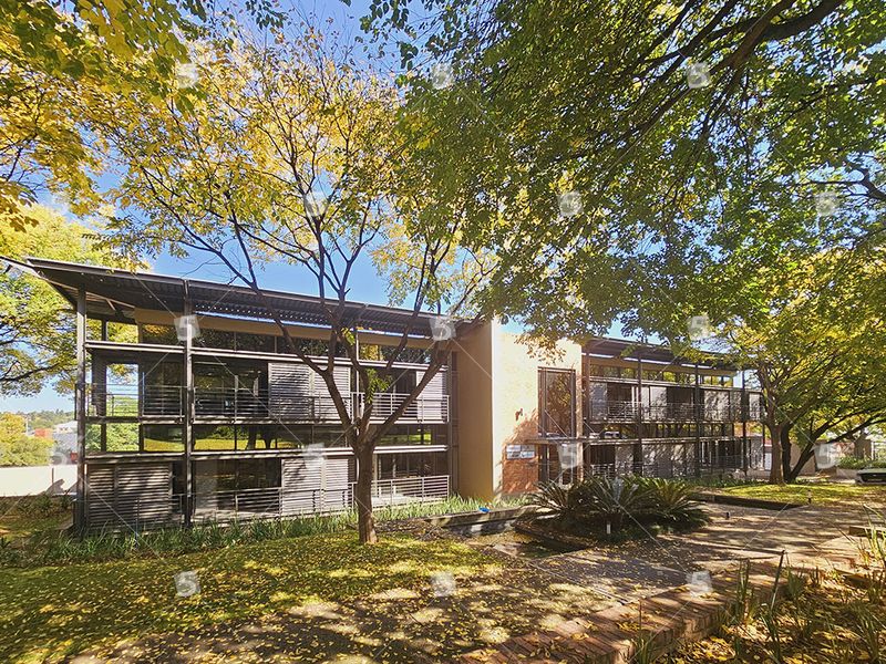 Modern multi-story office building featuring a steel and glass facade with integrated metal louvers, set within a mature landscaped garden environment.