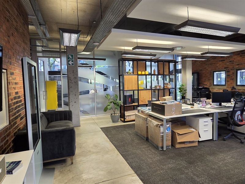 Modern open-plan office space featuring industrial design elements, including exposed brick walls, concrete ceilings, and a mix of carpeted and polished concrete flooring.