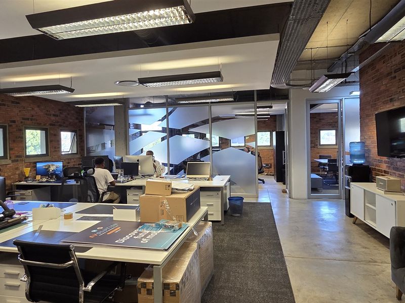 Modern open-plan office space featuring exposed brick walls, polished concrete floors, and industrial-style suspended lighting with glass partitioned meeting areas.