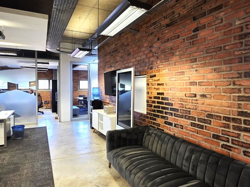 Modern open-plan office space featuring exposed brick walls, industrial-style exposed ceiling ducting, polished concrete floors, and glass-partitioned meeting rooms.
