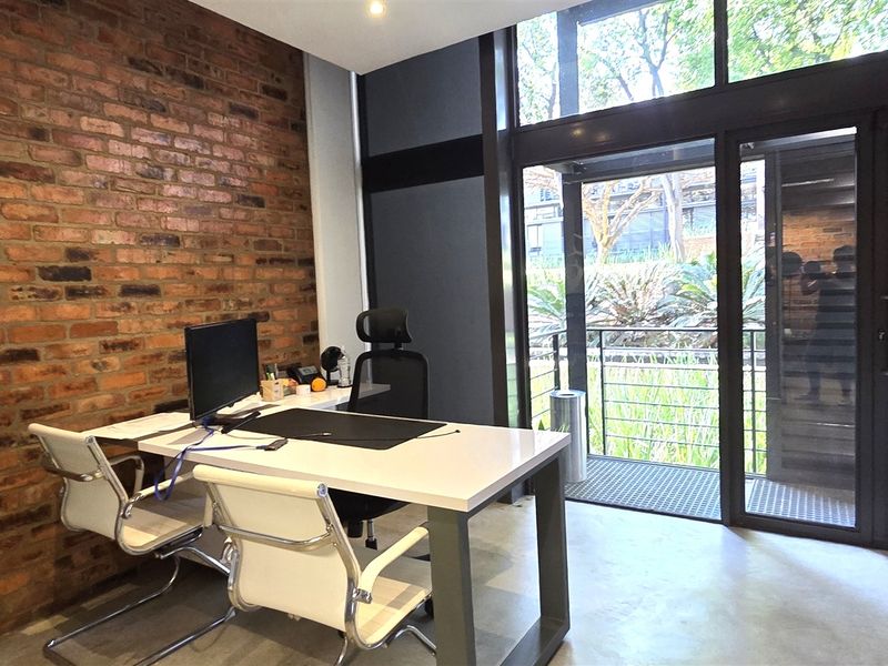 Modern office space featuring an exposed brick feature wall, polished concrete floors, and large floor-to-ceiling windows providing natural light and access to a balcony.
