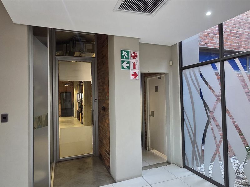 A modern office entrance area featuring glass doors, exposed brick accents, and clean tiled flooring with integrated safety signage and recessed lighting.