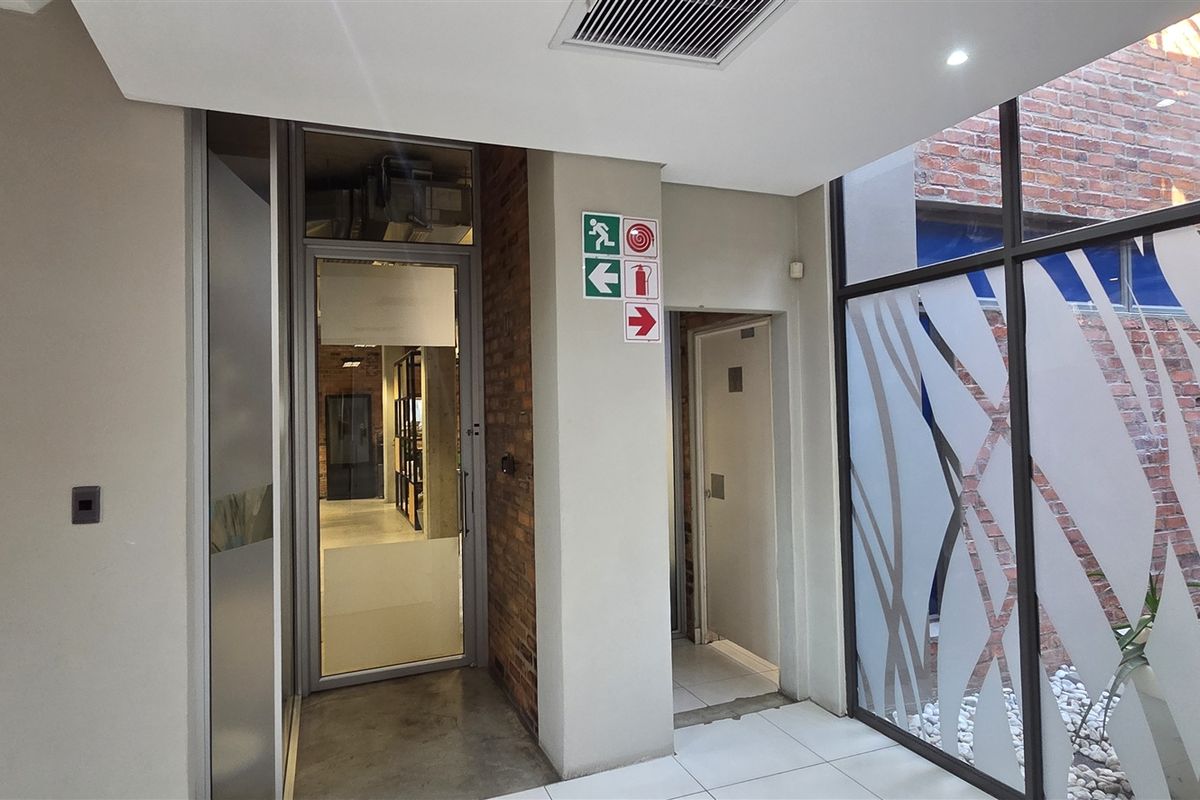 A modern office entrance area featuring glass doors, exposed brick accents, and clean tiled flooring with integrated safety signage and recessed lighting.