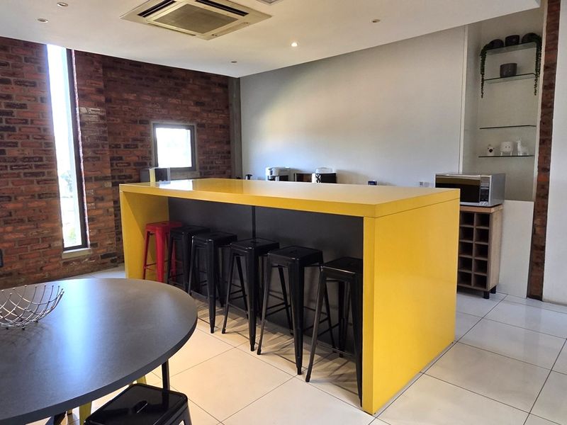 Modern office kitchen area featuring a prominent yellow central island, exposed brick feature wall, and sleek tiled flooring.