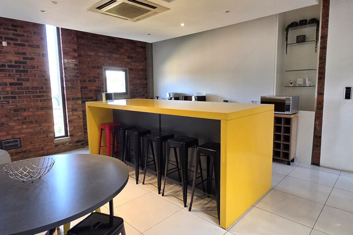 Modern office kitchen area featuring a prominent yellow central island, exposed brick feature wall, and sleek tiled flooring.
