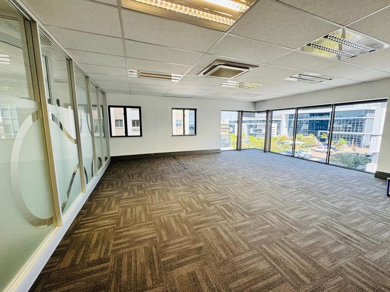 A bright, open-plan office space featuring carpet tiles, a suspended ceiling with recessed lighting, glass partitions, and large windows offering natural light.