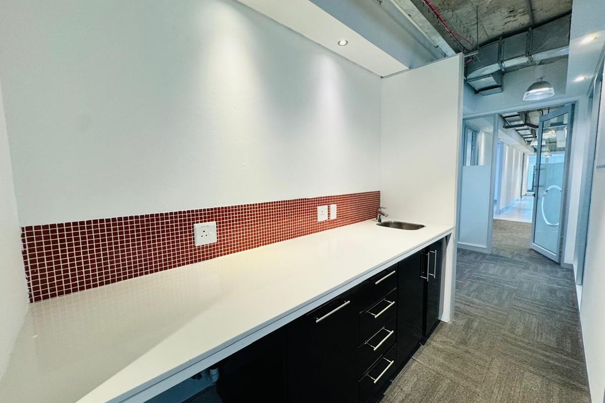 A modern office kitchenette featuring sleek black cabinetry, a white countertop, a small sink, and a red tiled backsplash with exposed industrial ceiling details.