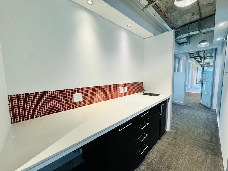 A modern office kitchenette featuring sleek black cabinetry, a white countertop, a small sink, and a red tiled backsplash with exposed industrial ceiling details.