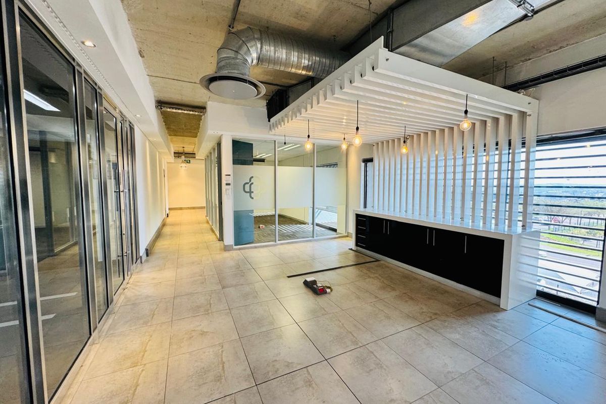 Modern open-plan office space featuring exposed industrial ceilings, glass partitions, tiled flooring, and a custom built-in reception or kitchenette area with slatted timber detailing.