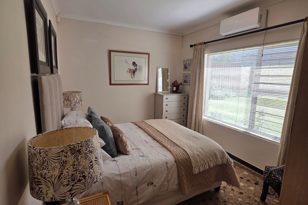 A cozy bedroom featuring a wall-mounted air conditioning unit, neutral wall finishes, and a window with fitted blinds.