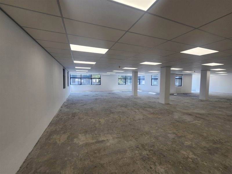 A spacious, open-plan office area featuring a suspended ceiling with recessed lighting, structural columns, and large windows providing ample natural light.
