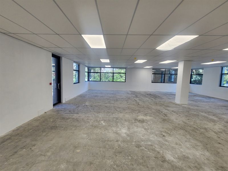 A spacious, open-plan office area featuring a suspended ceiling, recessed lighting, and large windows, currently awaiting final flooring installation.