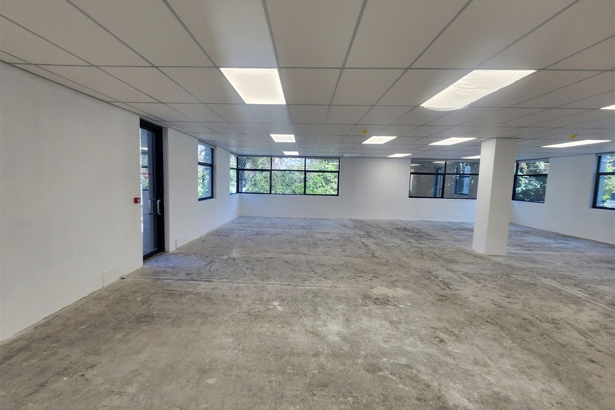 A spacious, open-plan office area featuring a suspended ceiling, recessed lighting, and large windows, currently awaiting final flooring installation.