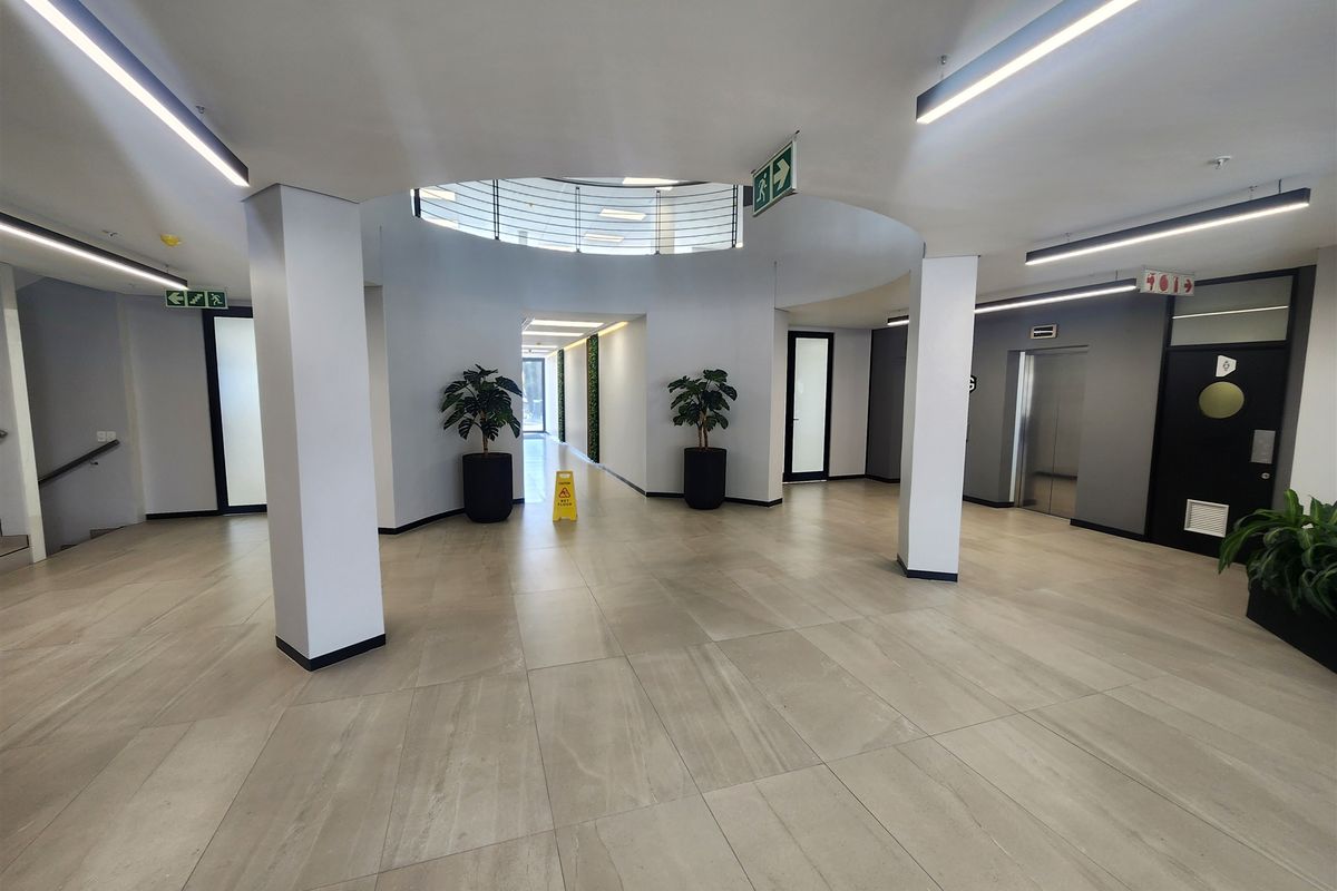 Modern office building lobby featuring tiled flooring, recessed linear lighting, a central circular ceiling feature, and elevator access.