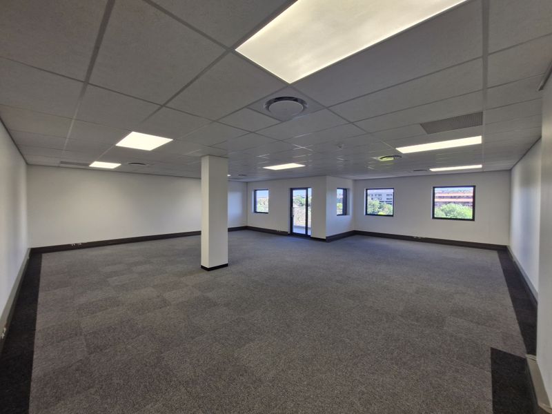 A spacious, empty open-plan office area featuring a suspended ceiling, recessed lighting, and carpeted flooring with large windows providing natural light.