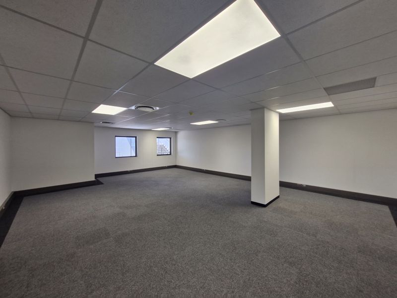 A spacious, empty commercial office area featuring neutral carpeting, a suspended ceiling with recessed lighting, and a central structural column.