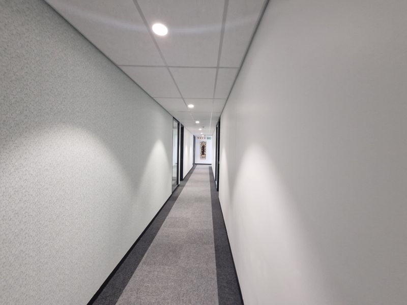 A clean, well-lit commercial office hallway featuring neutral carpet tiles, suspended ceiling grid, and glass partitions leading to private office spaces.