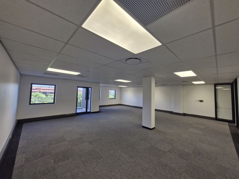 A spacious, open-plan commercial office area featuring neutral carpet tiles, a suspended ceiling with recessed lighting, and large windows providing natural light.