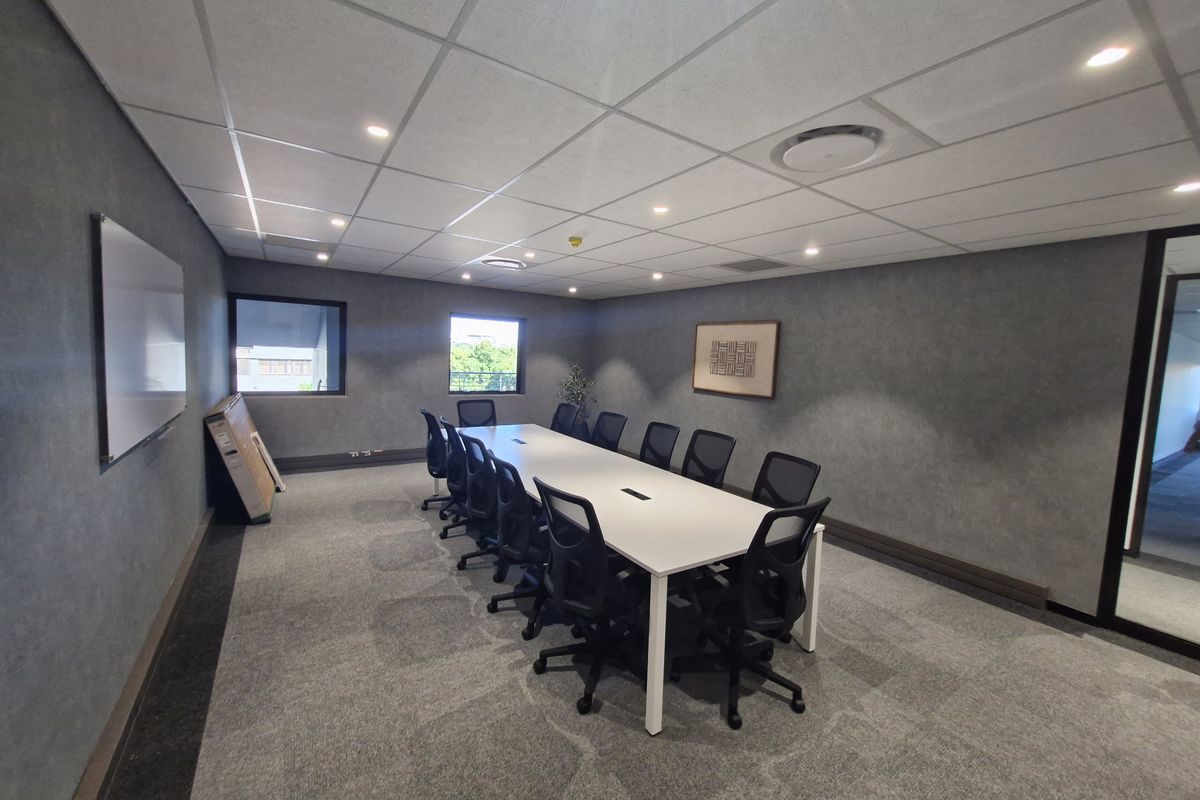 A professional boardroom or meeting room featuring a large central table, carpeted flooring, suspended ceiling with recessed lighting, and a wall-mounted whiteboard.