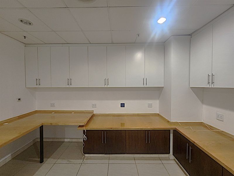 A clean, empty office space featuring extensive built-in cabinetry, laminate work surfaces, tiled flooring, and a suspended ceiling with recessed lighting.