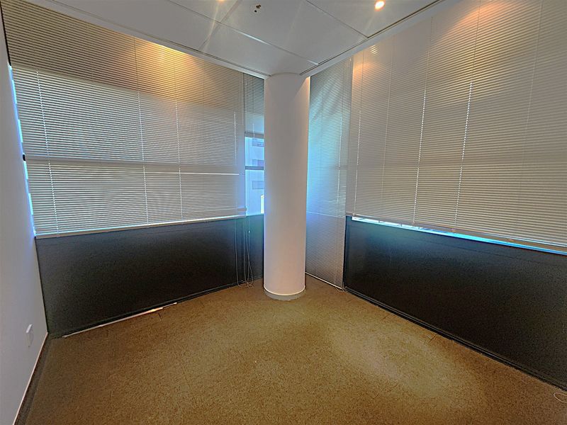 An empty office space featuring a central structural column, carpeted flooring, suspended ceiling with recessed lighting, and large windows with fitted venetian blinds.