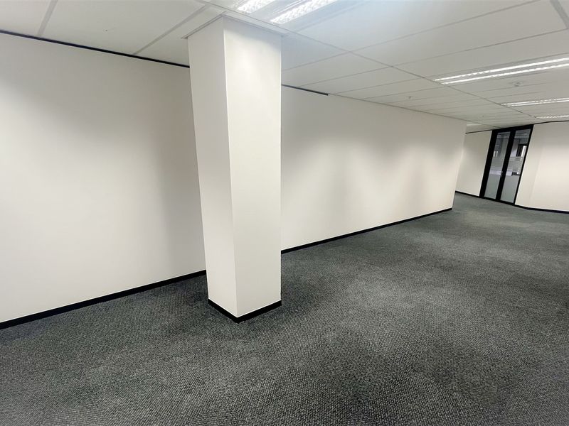 A spacious, empty open-plan office area featuring neutral walls, carpeted flooring, a structural column, and recessed ceiling lighting with a glass partition entrance.