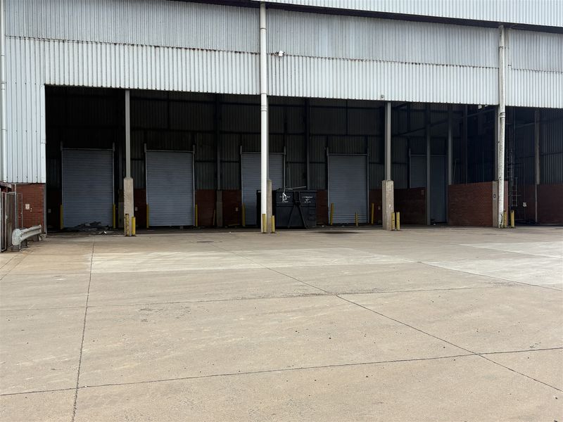 Industrial loading bay area featuring multiple roller shutter doors, concrete apron, and corrugated metal cladding structure.