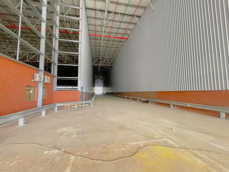 Spacious industrial warehouse area featuring high ceilings with exposed steel trusses, corrugated metal cladding, brick walls, and durable concrete flooring with safety crash barriers.