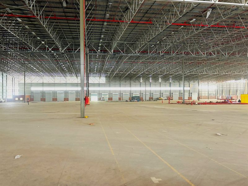 Expansive industrial warehouse space featuring high ceilings with exposed steel trusses, fire sprinkler systems, concrete flooring, and multiple roller shutter access doors.