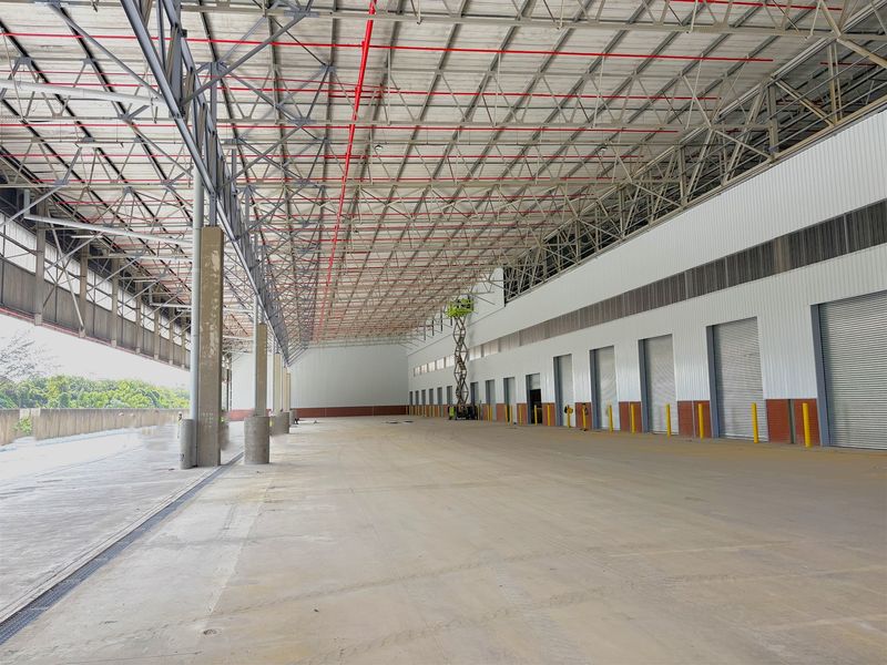 Spacious industrial warehouse interior featuring high ceilings with exposed steel trusses, fire sprinkler systems, concrete flooring, and multiple roller shutter loading doors.