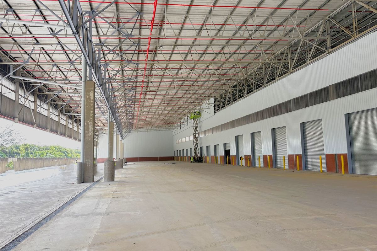 Spacious industrial warehouse interior featuring high ceilings with exposed steel trusses, fire sprinkler systems, concrete flooring, and multiple roller shutter loading doors.