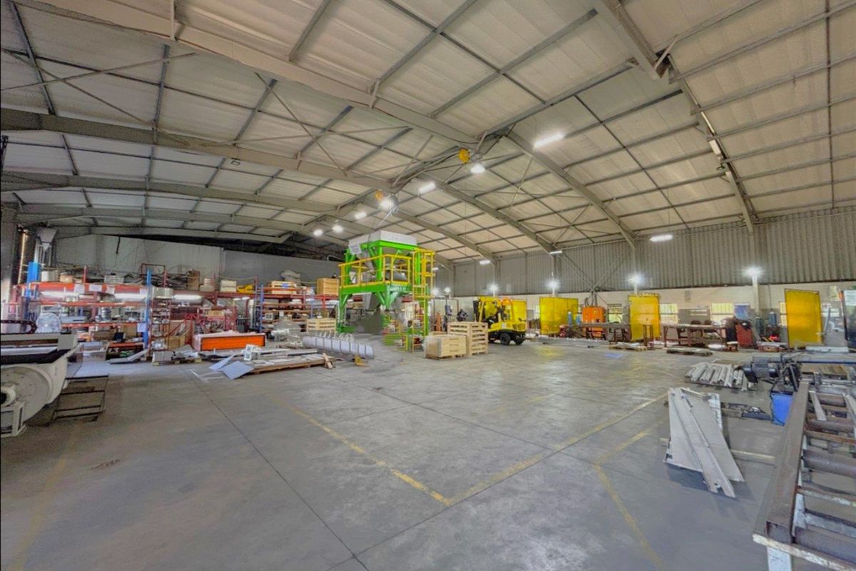 Spacious industrial warehouse facility featuring high ceilings with exposed steel trusses, concrete flooring, and functional industrial lighting throughout the open workspace.